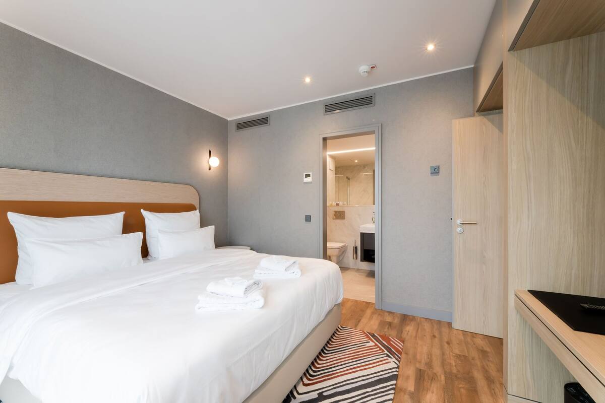 A modern hotel room with a large bed, a wooden wardrobe, and a bathroom visible through an open door.