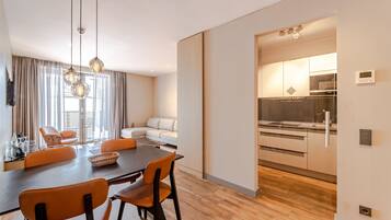 A modern dining area with a wooden table and orange chairs, a sofa, and a kitchen in the background.
