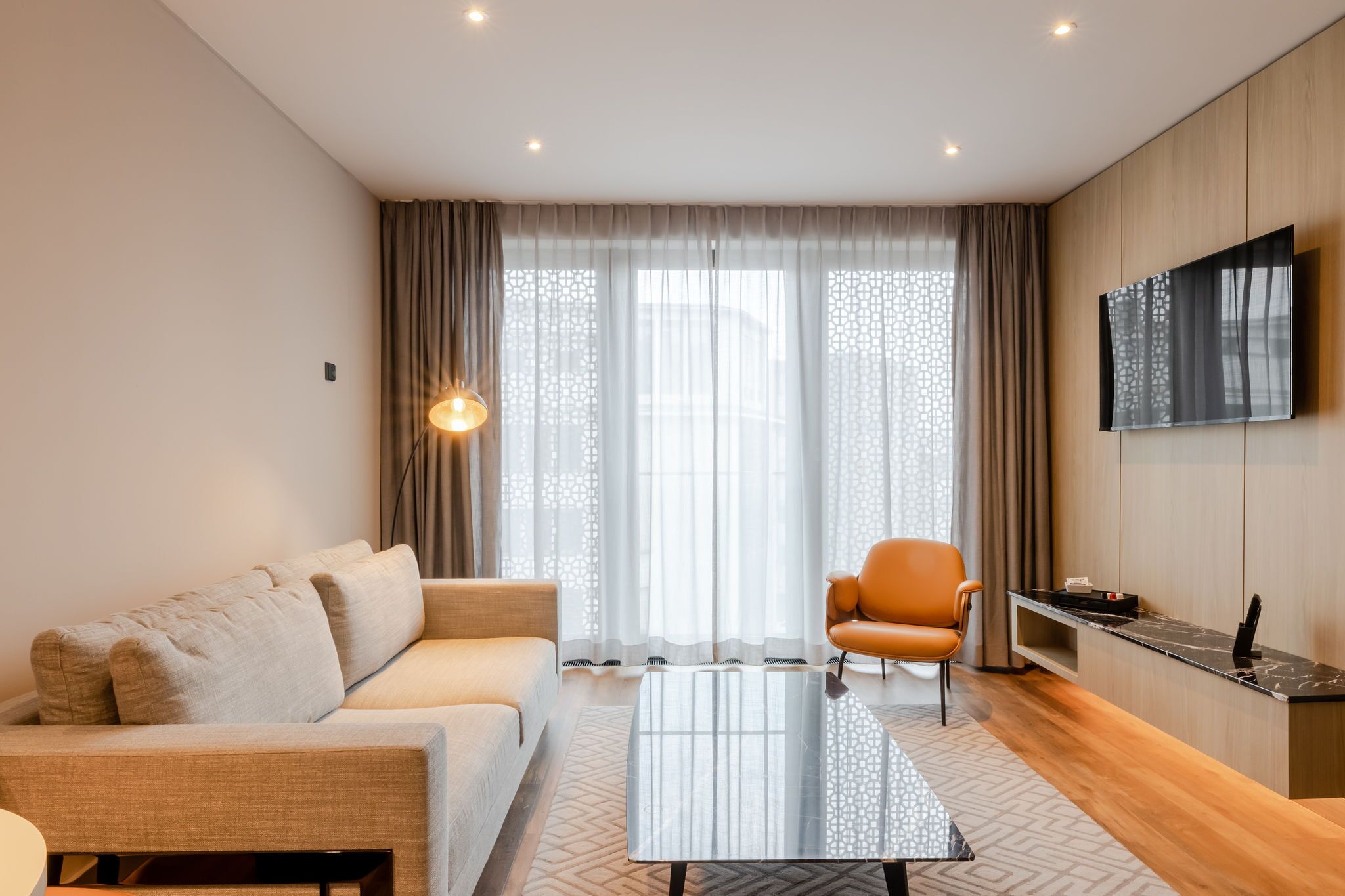 A modern living room with a sofa, a coffee table, a TV mounted on the wall, and a window with sheer curtains.