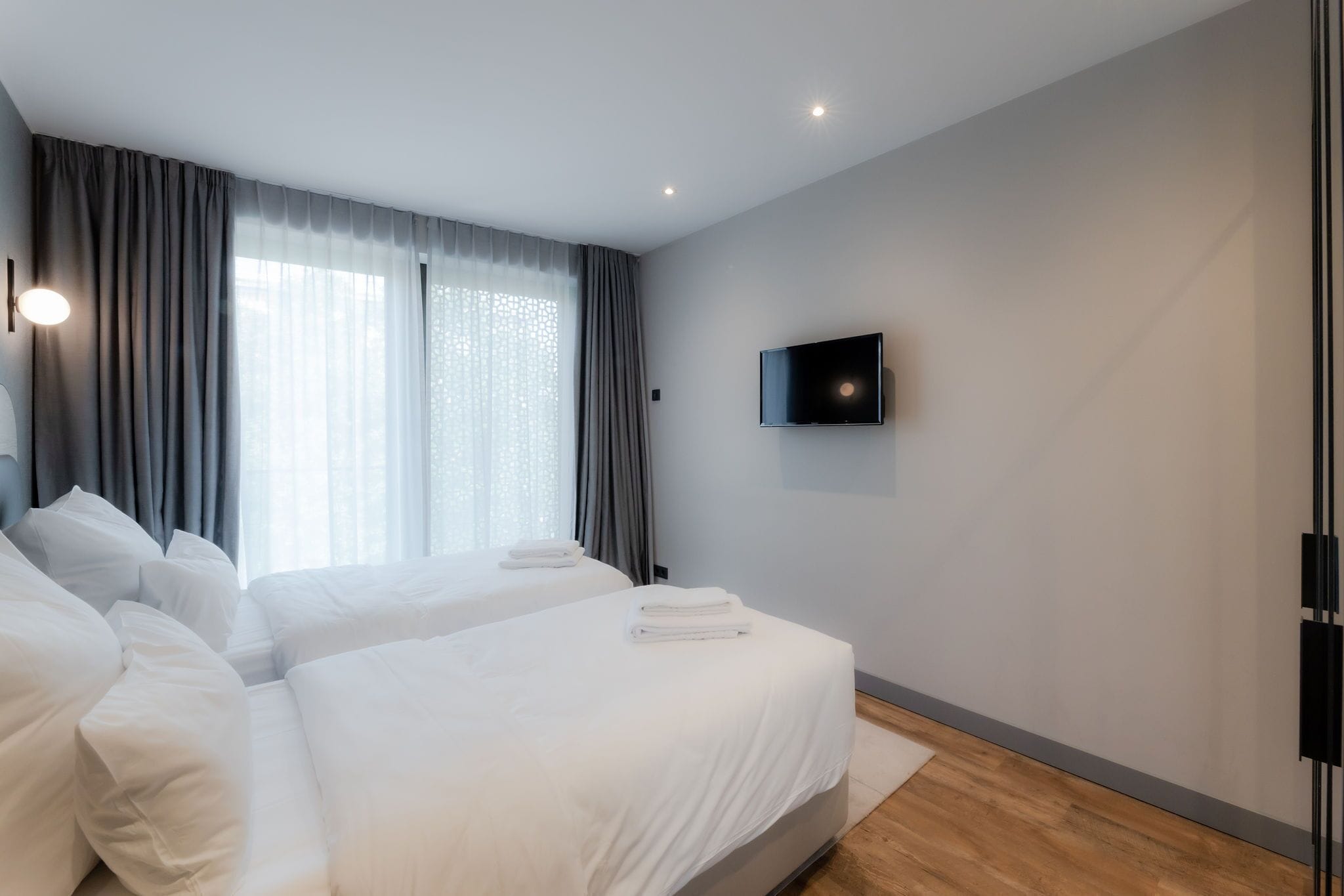 A modern hotel room with two beds, a flat-screen TV mounted on the wall, and large windows with grey curtains.