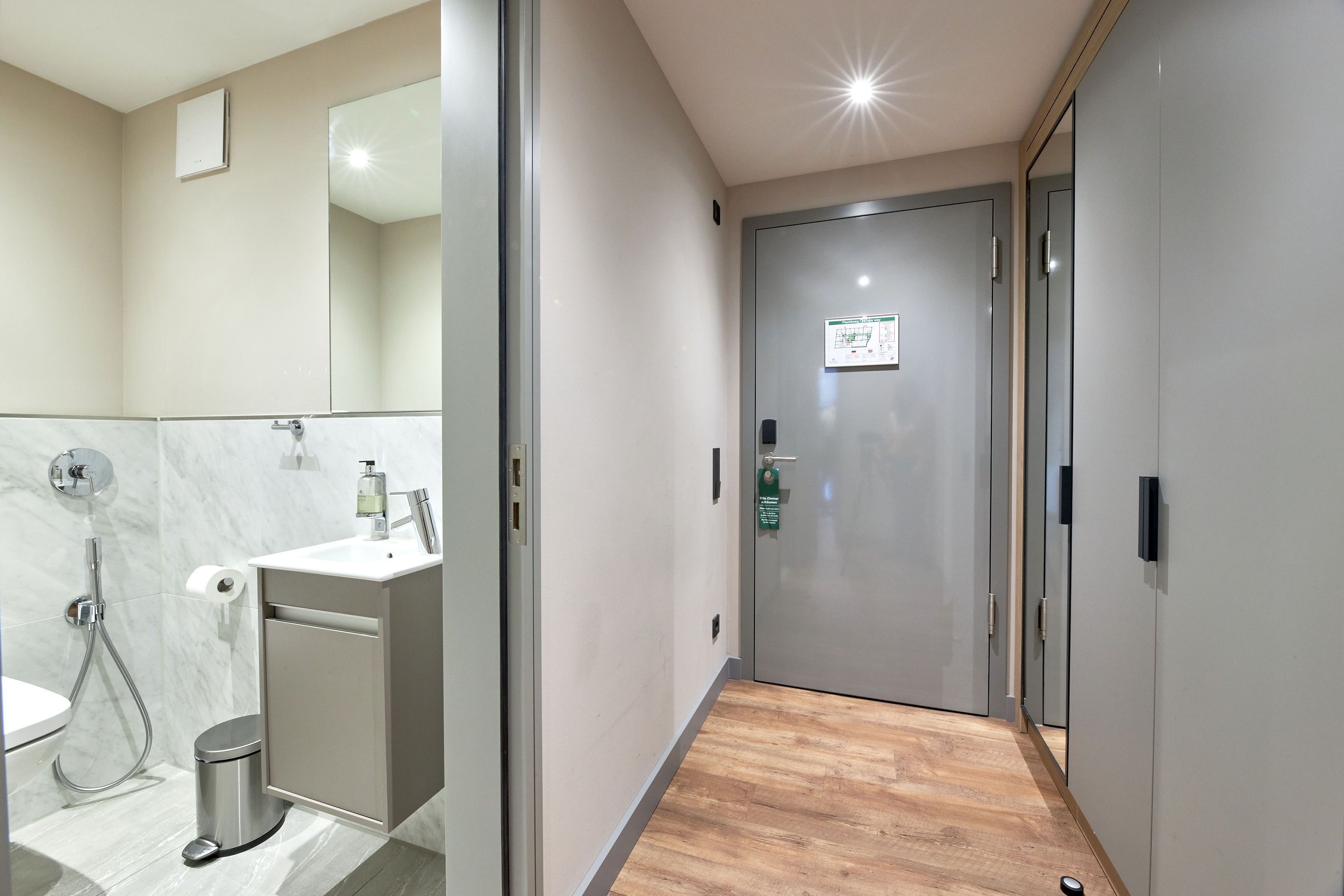 A modern bathroom with a shower, toilet, and sink, and a hallway leading to a door with a sign.