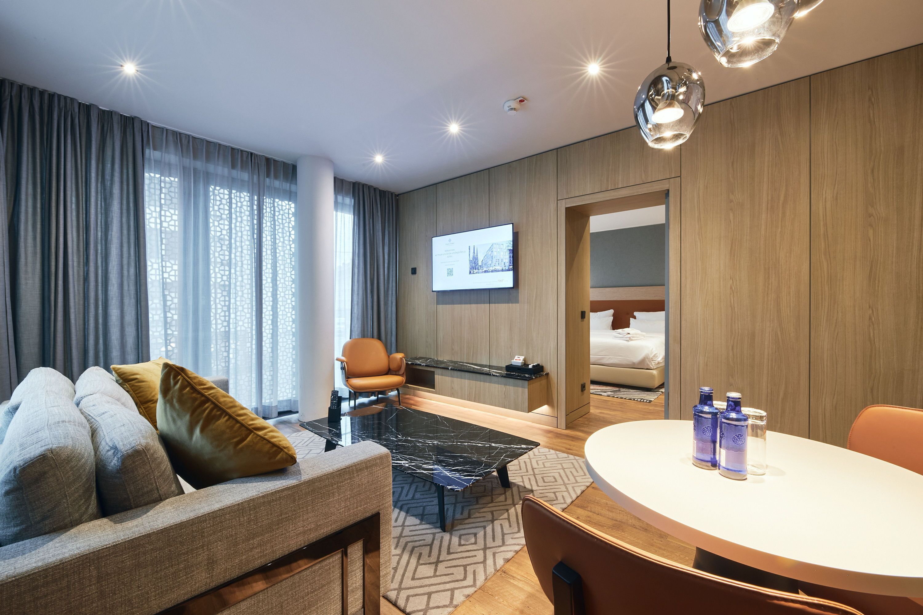 Une chambre d’hôtel moderne équipée d’un canapé, d’une table à manger, d’un téléviseur fixé au mur et d’une armoire en bois.