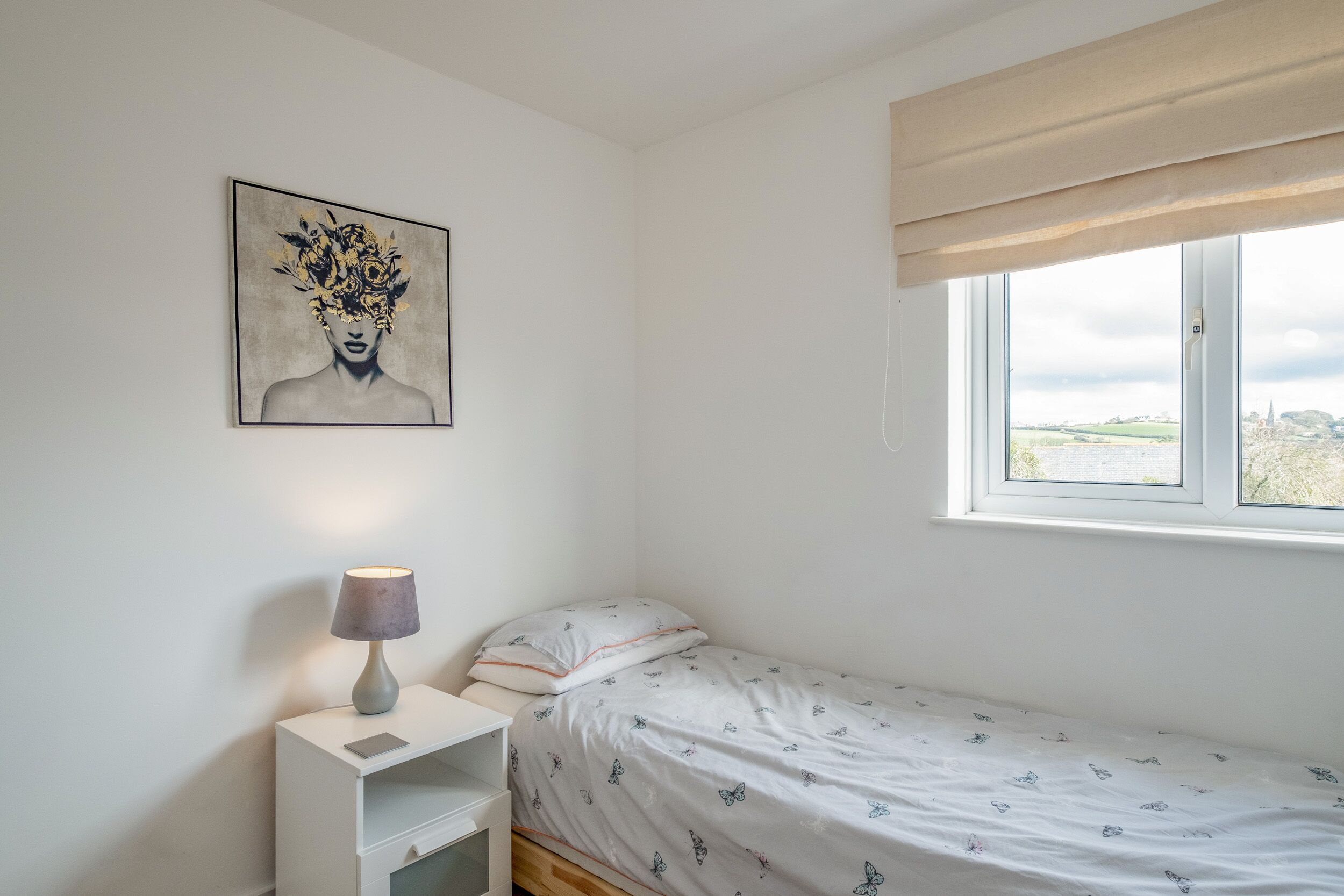 A bedroom with a bed, a bedside table with a lamp, a window with a view, and a painting on the wall.