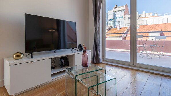 A modern living room with a flat-screen TV on a white cabinet, a glass coffee table, and a view of buildings through a large window.