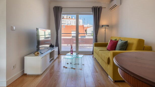 Een moderne woonkamer met een bank, een televisie, een glazen salontafel en een uitzicht op gebouwen door grote ramen.