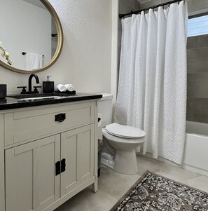 Une salle de bain avec un lavabo blanc, un robinet noir et des toilettes. On y trouve un rideau de douche et un tapis à motifs sur le sol.