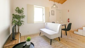 A modern living room with a sofa, a dining table with chairs, a potted plant, a remote control, and a window with blinds.