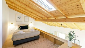 Ein Schlafzimmer mit schrägem Dach, Holzbalken, einem Bett mit Kissen, einem Nachttisch, einer Pflanze und gerahmten Bildern an der Wand.
