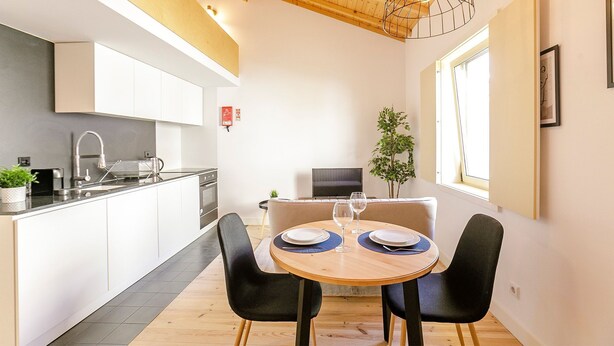 Eine moderne Küche mit weißen Schränken, einer schwarzen Arbeitsplatte und einem eingebauten Backofen. Es gibt einen Essbereich mit einem runden Holztisch, zwei schwarzen Stühlen und einem kleinen Topfpflanzen.