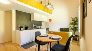 A compact kitchen with white cabinets, a black countertop, and a yellow accent wall. There is a dining table with two chairs and a small potted plant.