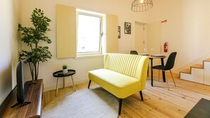 A modern living room with a yellow sofa, a wooden coffee table, a small side table with a plant, a television on a wooden stand, a window with a view, and a staircase leading to another room.