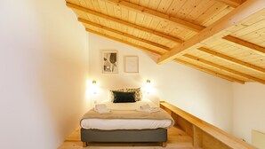 A bedroom with a sloped ceiling, wooden beams, a bed with pillows, and two wall-mounted lights.