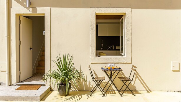 A small outdoor seating area with a table and chairs, a potted plant, and a window with a view of the interior.