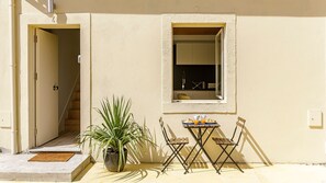 A small outdoor seating area with a table and chairs, a potted plant, and a window with a view of the interior.