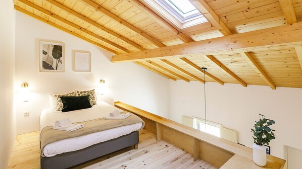 A bedroom with a sloped ceiling, wooden beams, a bed with a dark headboard, a bedside table, a nightstand, a wall-mounted shelf, and a potted plant.