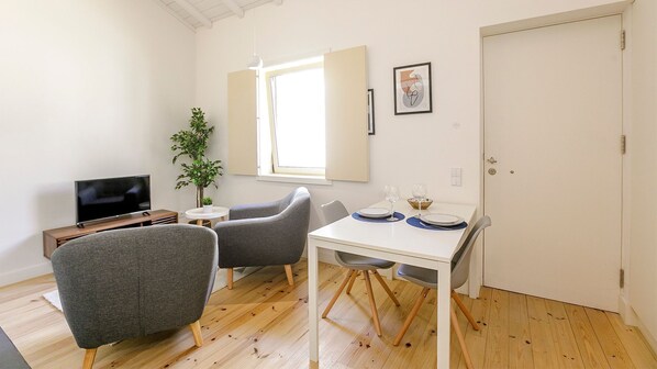 A small, bright room with a TV, two grey chairs, a white table with plates and glasses, a potted plant, and a window with blinds.