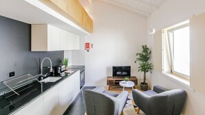 Um espaço de estar compacto com cozinha compacta, cadeiras estofadas em cinza, uma pequena mesa redonda, uma televisão e um vaso com planta.
