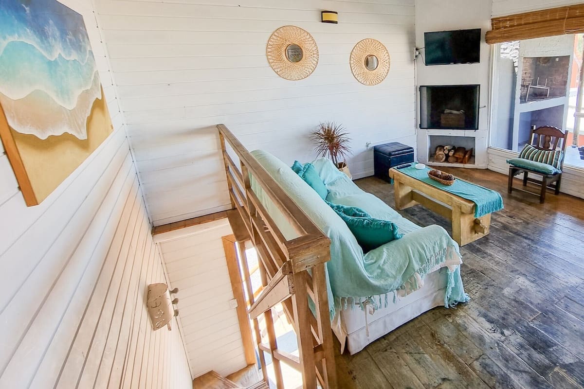 Ett mysigt rum med en trätrappa, en vit soffa med turkosa kuddar, ett träbord och en väggmonterad TV.