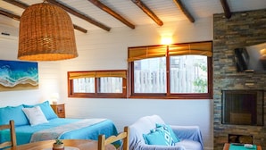 A cozy room with a wooden ceiling, a stone fireplace, a large window with curtains, a bed with blue bedding, and a dining table with a placemat and a small plant. - Cabañas Kundalini Punta de Diablo (Punta del Diablo)