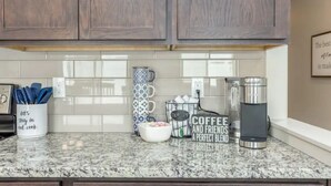 Une cuisine avec des meubles en bois, des carreaux de céramique blancs disposés en motif métro, un plan de travail en granit, une machine à café, un porte-tasses et un saladier rempli de fruits.