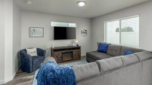 Un salon avec un canapé gris, un fauteuil bleu, un téléviseur à écran plat fixé au mur et une cheminée.