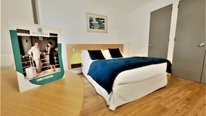 A hotel room with a bed, a nightstand, a lamp, a framed picture on the wall, and a door. - hotel thermes les Ecureuils (Dax)