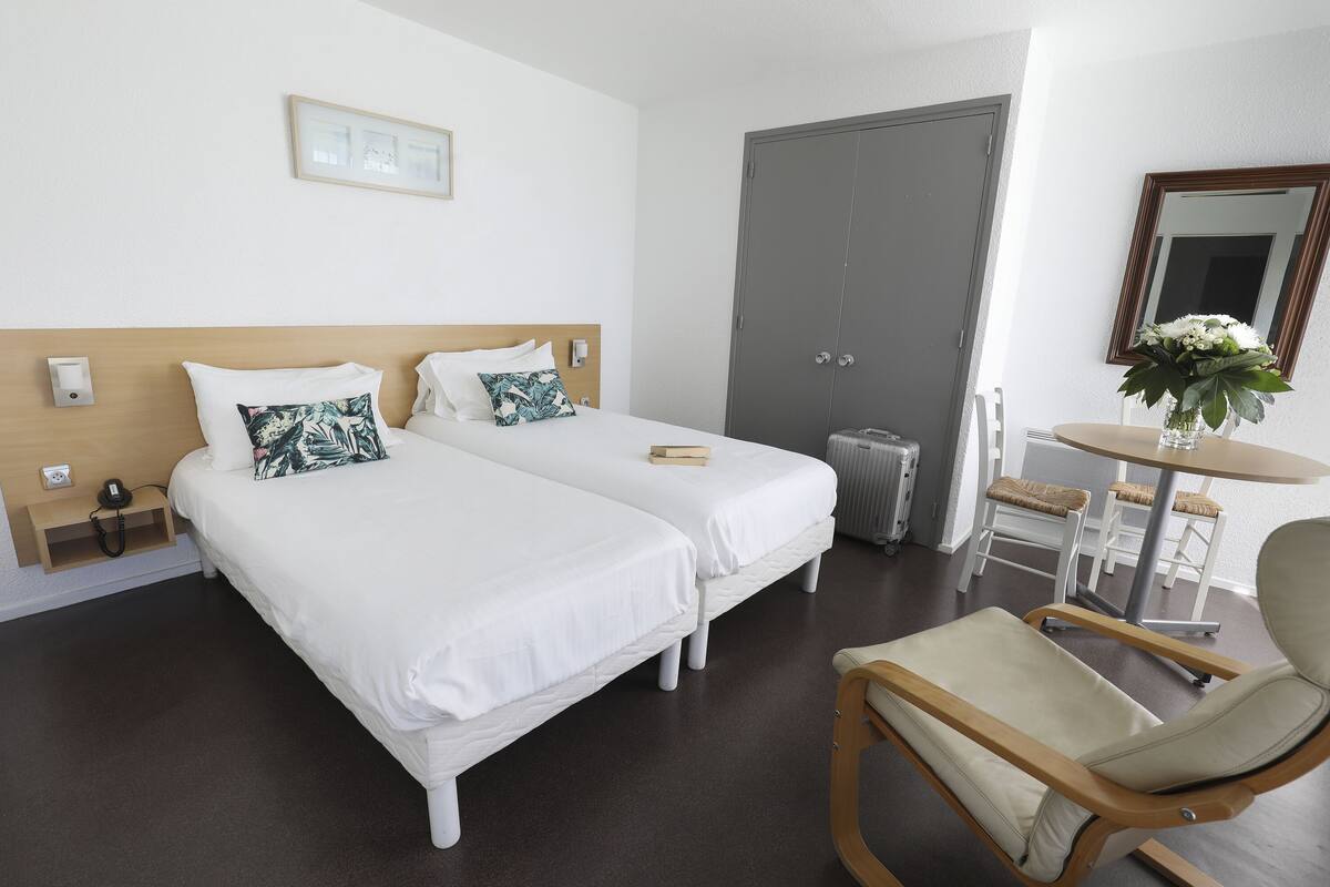 Een hotelkamer met twee eenpersoonsbedden, een houten hoofdeinde, een klein tafeltje met een bloevas, een stoel en een grijze deur.