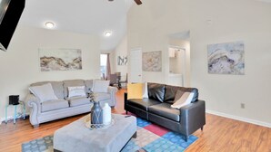 Un salon spacieux avec un canapé gris, un canapé noir et une table basse. On y trouve des ventilateurs de plafond, un éclairage encastré et des œuvres d’art accrochées aux murs.