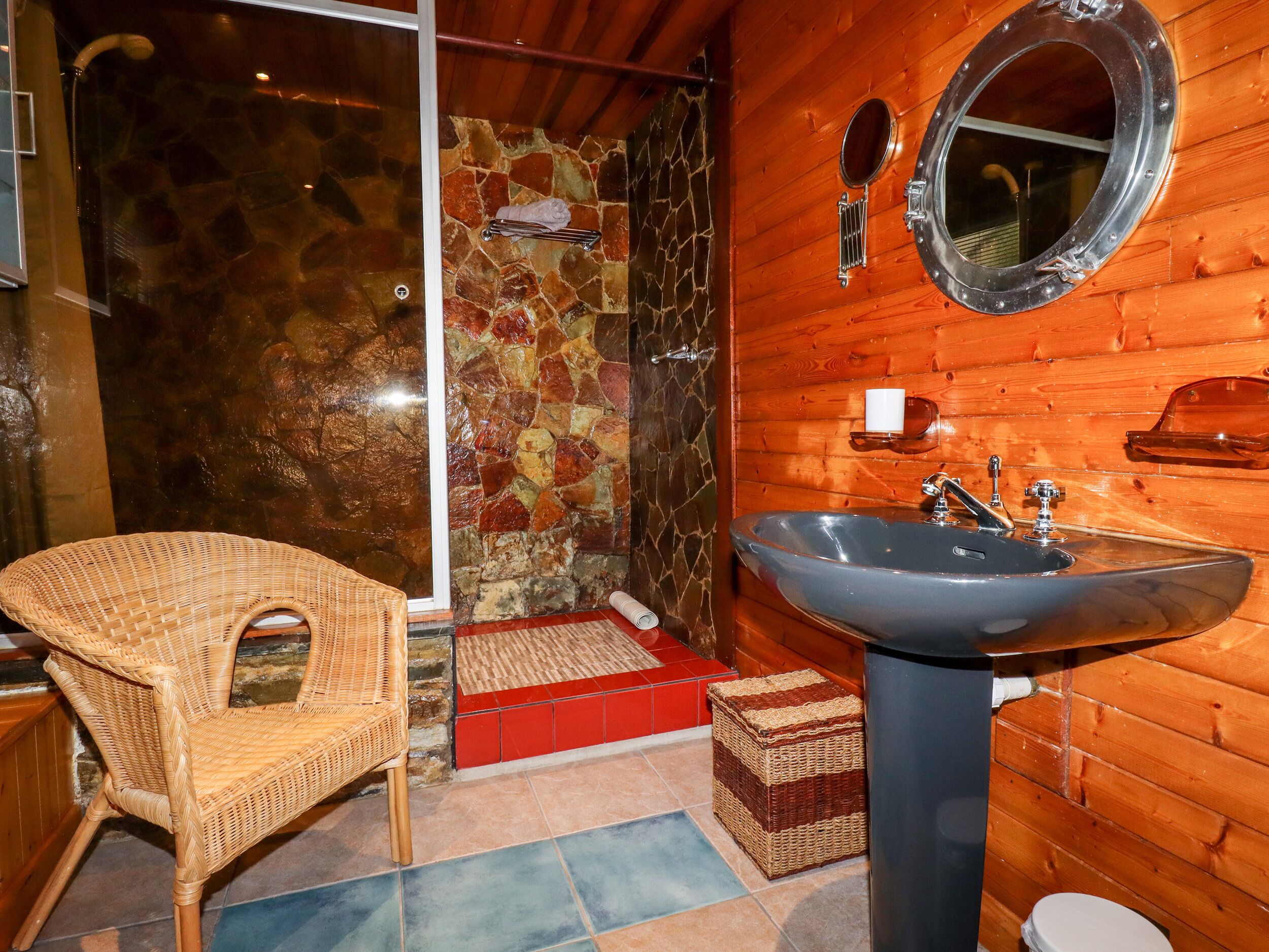 A bathroom with a stone wall, wooden paneling, a large sink, a mirror, a wicker chair, and a shower area.