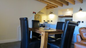 A dining area with a wooden table and black chairs, a kitchen in the background, and exposed wooden beams on the ceiling. - Farthing Cottage (Bishop Auckland)