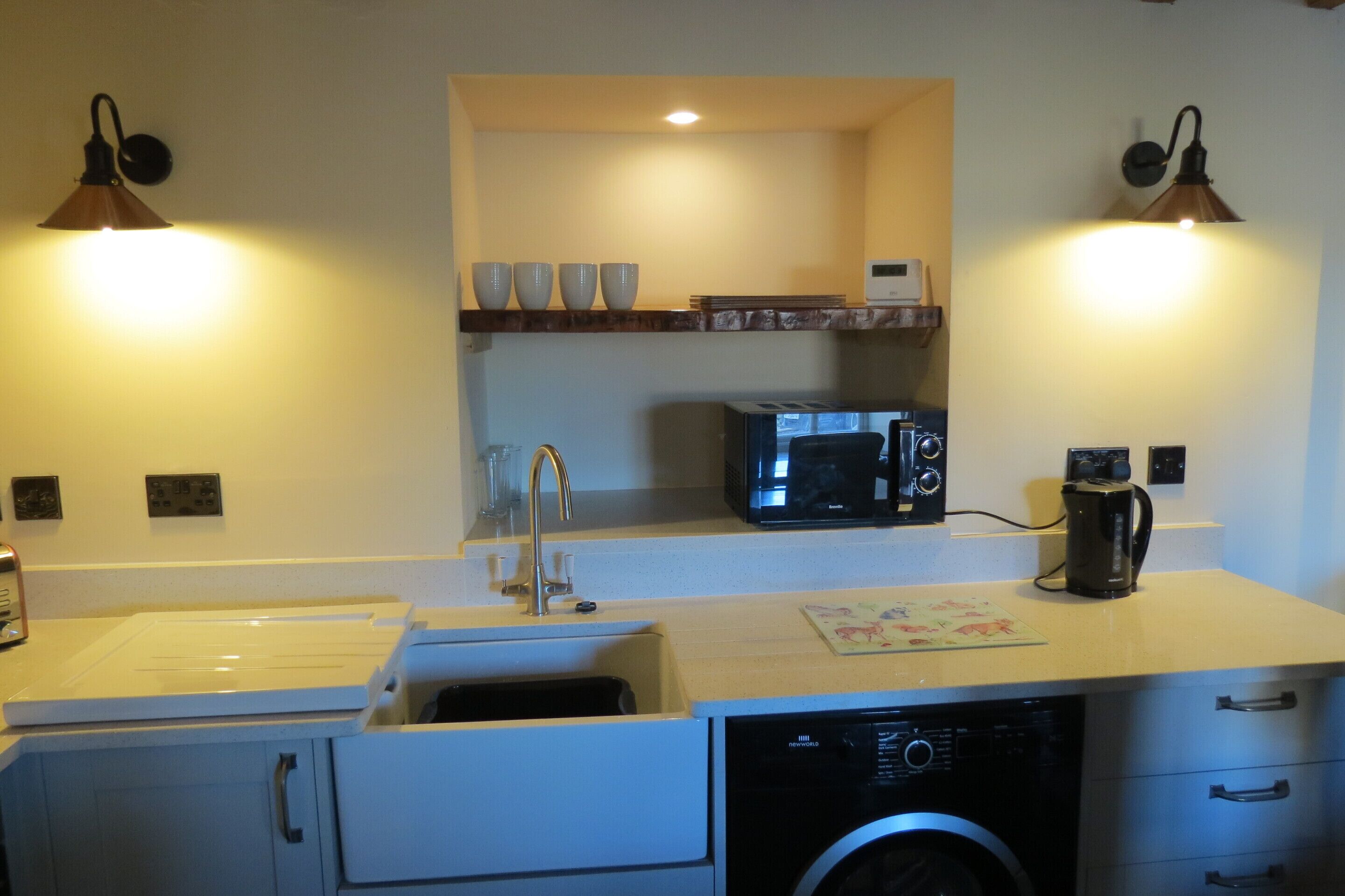 A compact kitchen with a microwave, coffee maker, and a sink. There are two wall-mounted lamps and a shelf with cups.