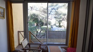 A room with a large sliding glass door leading to a backyard, a wooden chair, a small table, and a bed with a red bedspread. - Apartment (Saint-Mandrier-sur-Mer)