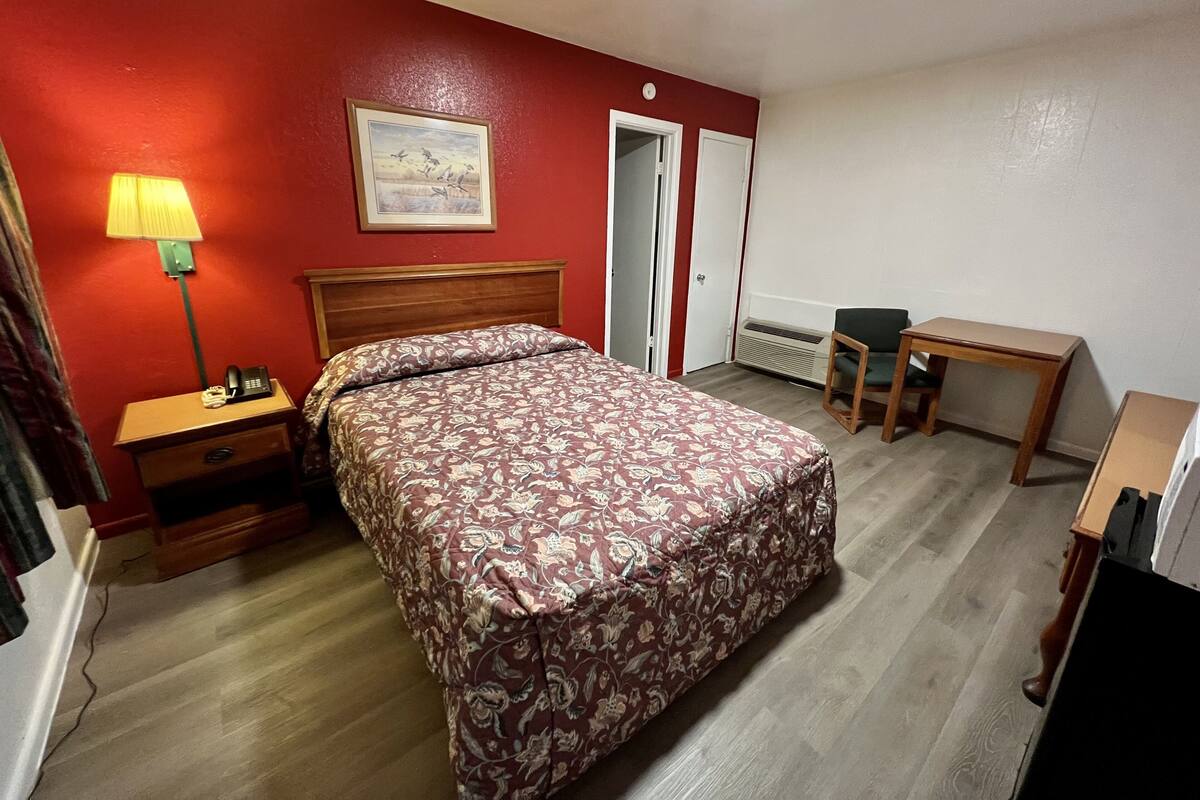 A hotel room with a bed, bedside table, lamp, desk, chair, and a radiator.