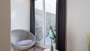 A room with a grey armchair, a small table with a bird sculpture, and a view of a stone wall through a glass door. - Peaceful & Sunny Building |One Terrace by HostWise (Porto)
