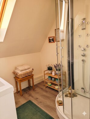 Un baño moderno con cabina de ducha de vidrio, suelo de madera, una mesita con toallas y un estante con artículos de aseo.