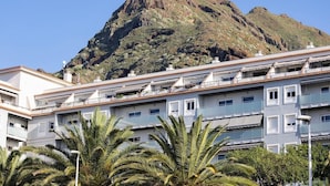 A multi-story building with balconies, a mountain in the background, and palm trees in the foreground.