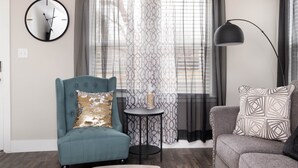 A living room with a teal armchair, a grey sofa, a round wall clock, a floor lamp, and a patterned curtain.
