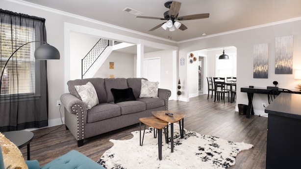 A modern living room with a gray sofa, a blue armchair, and a wooden coffee table. There is a staircase leading to another level, a dining area with chairs, and a kitchen area with a black countertop.
