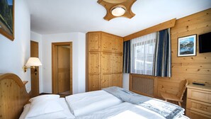A bedroom with a wooden headboard, a bed with white linens, a wooden wardrobe, a desk with a chair, and a window with curtains.
