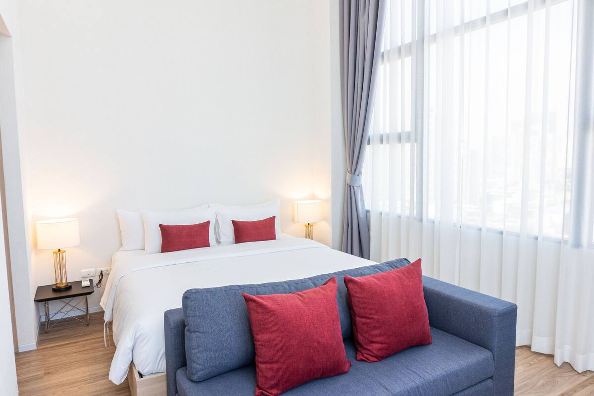 A hotel room with a bed, a sofa, a nightstand with a lamp, and a window with curtains.