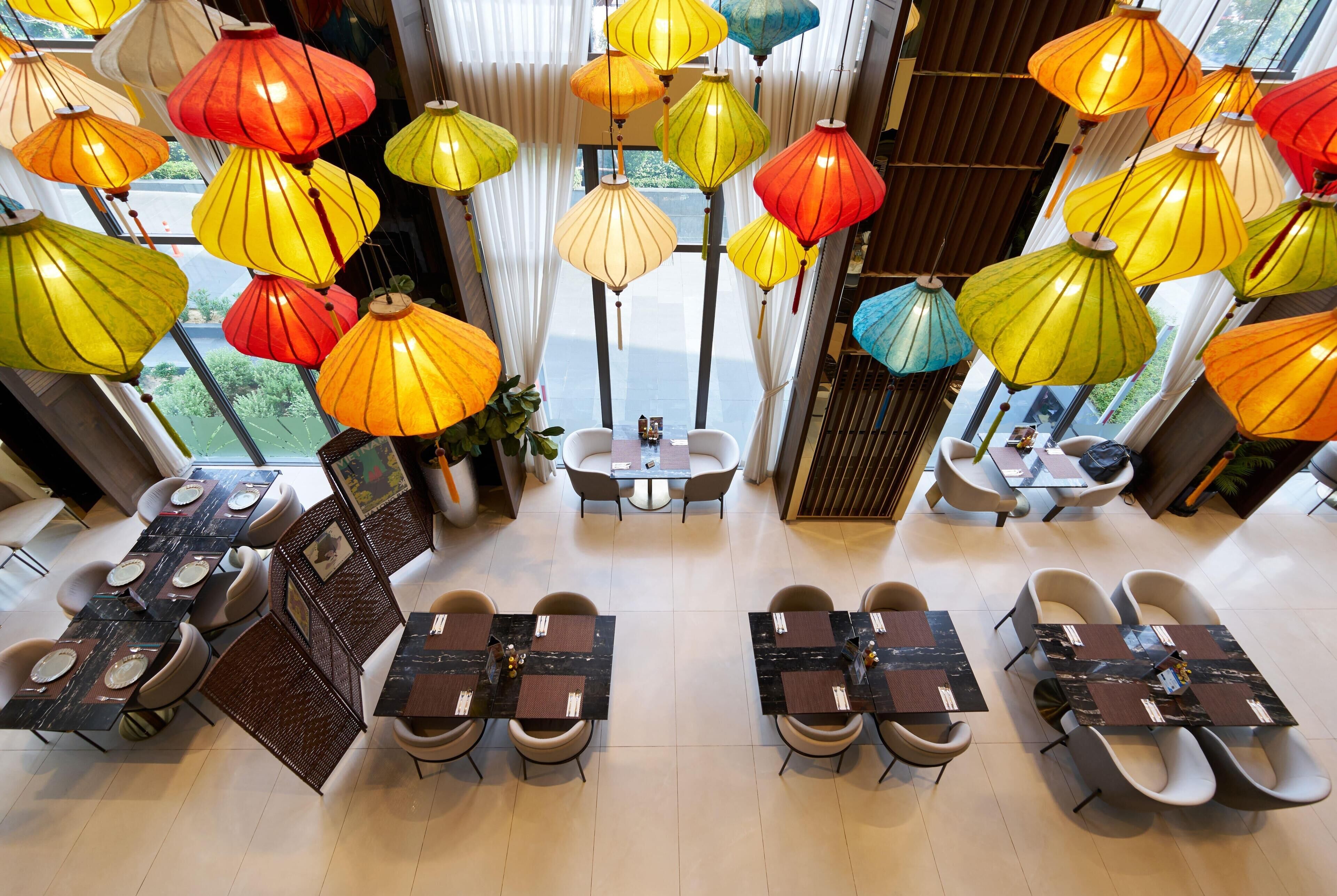 A restaurant with multiple tables and chairs, colorful hanging lanterns, and a view through large windows.