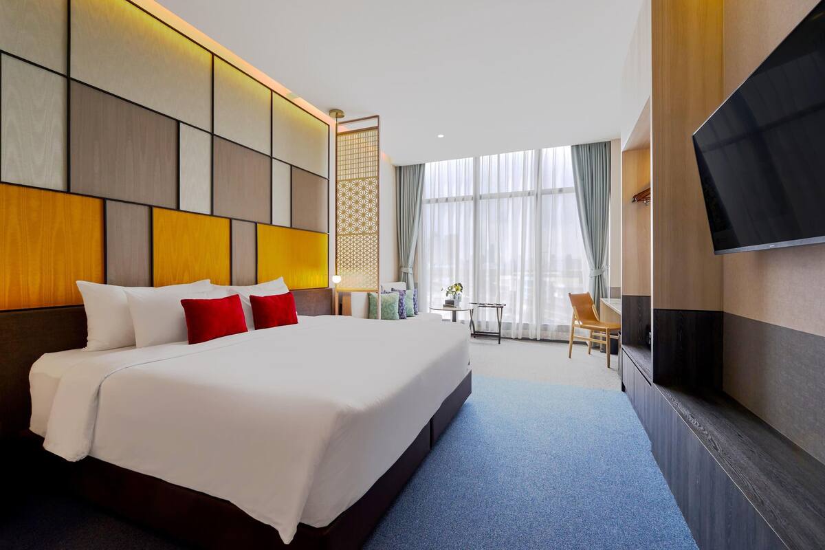 A modern hotel room with a large bed, a flat-screen TV, and a view of the city through large windows.