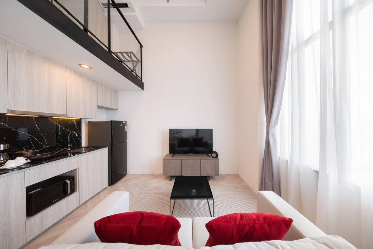 A modern living room with a sofa, red cushions, a coffee table, a TV, and a kitchen area with a stove and oven.