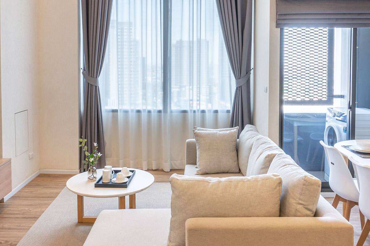 A modern living room with a sectional sofa, a round coffee table, and a view of a cityscape through large windows with curtains.