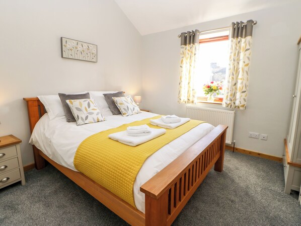 A neatly made bed with a yellow blanket and pillows, a wooden headboard, a bedside table, and a window with curtains.