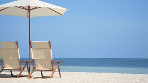 Deux chaises de plage et un parasol blanc sur une plage de sable, avec un océan bleu clair en arrière-plan.