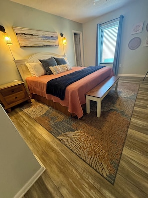 Un dormitorio con cama, mesitas de noche, un banco, una alfombra y un cuadro en la pared.
