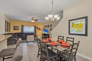 A modern living room with a sofa, a dining table set with orange napkins, and a TV in the background.