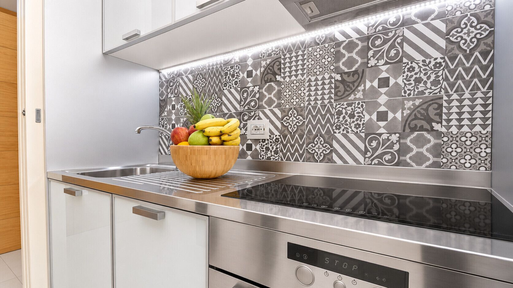 Une cuisine moderne avec un évier en acier inoxydable, un panier de fruits posé sur le plan de travail et des carreaux décoratifs sur la crédence.