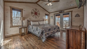 A cozy bedroom with a large bed, wooden floors, a ceiling fan, and a window with a view of trees. - Wolfs Den II - Shuffleboard | Foosball | Fire Pit | Outdoor Wood Burning Fireplace (Blue Ridge)
